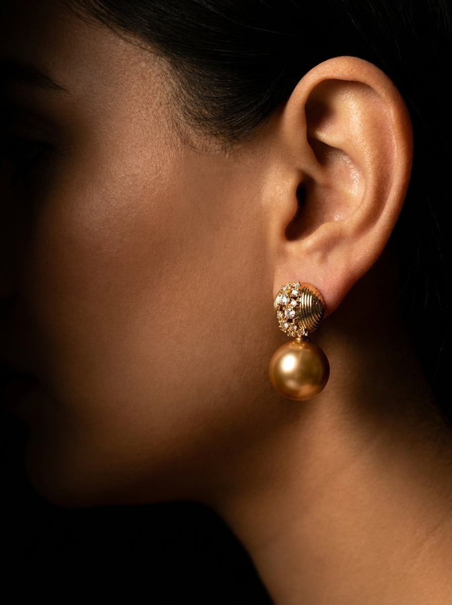A close-up side profile of a woman's ear wearing a gold drop earring with a textured, diamond-like top and a large round pearl pendant, set against a dark background.
