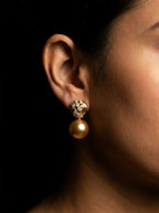 A close-up of a model's ear wearing a gold drop earring with a small ornate stud and a round pearl-like drop, set against a dark background.