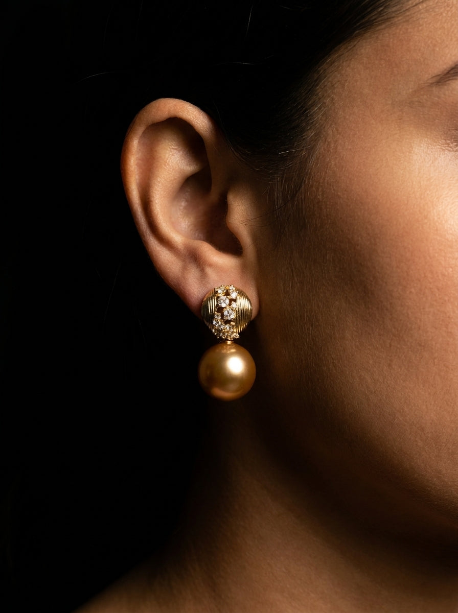 A close-up of a model's ear wearing a gold drop earring with a small ornate stud and a round pearl-like drop, set against a dark background.